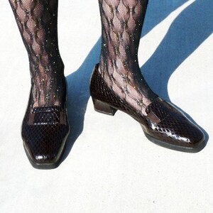 Op de afbeelding: Een paar donkerbruine, glanzende leren loafers met een slangenpatroon. De schoenen hebben een vierkante neus en een kleine hak. De persoon draagt zwarte visnetkousen met glitter.