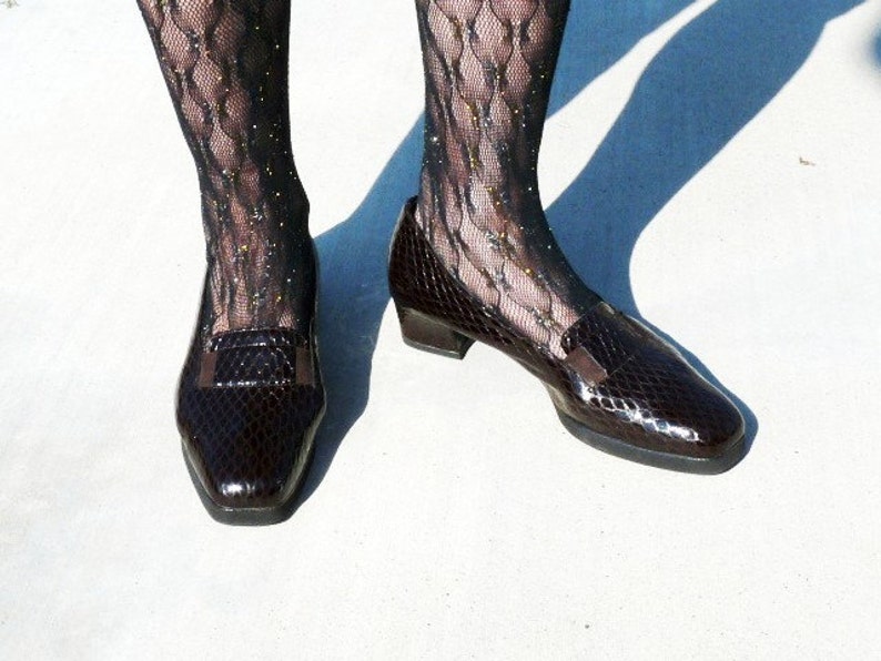 Op de afbeelding: Een paar donkerbruine, glanzende leren loafers met een slangenpatroon. De schoenen hebben een vierkante neus en een kleine hak. De persoon draagt zwarte visnetkousen met glitter.
