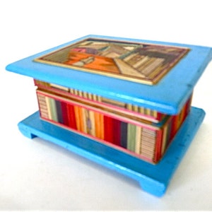 May include: A small, blue wooden box with a colorful, striped interior and a painted scene on the lid. The box has a hinged lid and a base with feet.