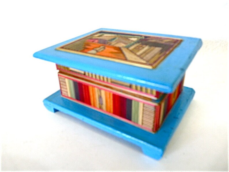 May include: A small, blue wooden box with a colorful, striped interior and a painted scene on the lid. The box has a hinged lid and a base with feet.