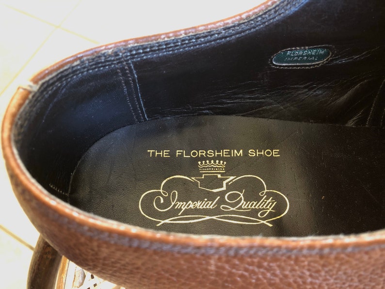 May include: Close-up of the inside of a brown leather shoe. The insole is black with gold lettering that reads "The Florsheim Shoe" and "Imperial Quality" with a crown design.