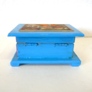 May include: A small, blue wooden box with a hinged lid. The top of the box is decorated with a colorful image. The box has a simple design and is perfect for storing small items.