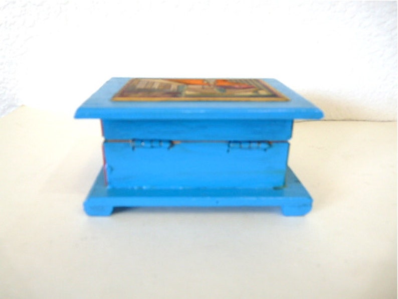 May include: A small, blue wooden box with a hinged lid. The top of the box is decorated with a colorful image. The box has a simple design and is perfect for storing small items.