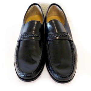 Pode incluir: Um par de sapatos de couro preto com um detalhe tran&ccedil;ado na frente. Os sapatos t&ecirc;m uma ponta pontiaguda e um acabamento polido.