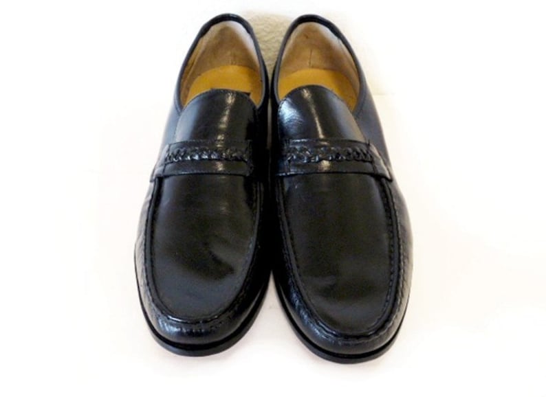 Pode incluir: Um par de sapatos de couro preto com um detalhe tran&ccedil;ado na frente. Os sapatos t&ecirc;m uma ponta pontiaguda e um acabamento polido.