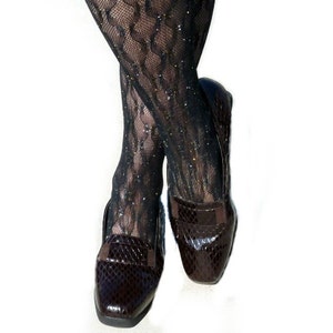 Op de afbeelding: Zwarte panty's met patroon en glitteraccenten worden gecombineerd met donkerbruine leren loafers. De loafers hebben een slangenleertextuur en een klein bandje. Witte achtergrond.