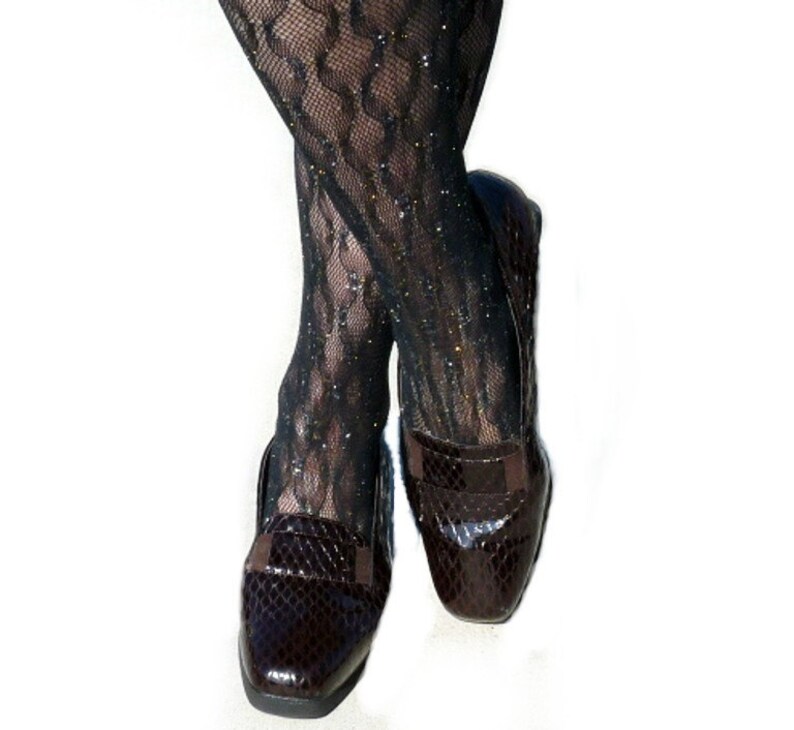 Op de afbeelding: Zwarte panty's met patroon en glitteraccenten worden gecombineerd met donkerbruine leren loafers. De loafers hebben een slangenleertextuur en een klein bandje. Witte achtergrond.