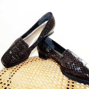 Op de afbeelding: Een paar bruine, glanzende loafers van imitatieleer met slangenprint. De schoenen hebben een vierkante neus, een kleine hak en een elastisch detail aan de bovenkant. De binnenkant is lichtbeige. De schoenen worden gepresenteerd op een lichtgekleurde rieten stoel.