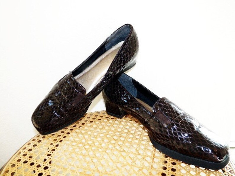 Op de afbeelding: Een paar bruine, glanzende loafers van imitatieleer met slangenprint. De schoenen hebben een vierkante neus, een kleine hak en een elastisch detail aan de bovenkant. De binnenkant is lichtbeige. De schoenen worden gepresenteerd op een lichtgekleurde rieten stoel.