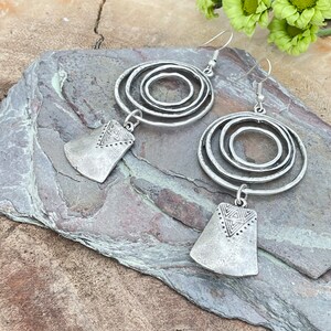 May include: A pair of silver-toned earrings. Each earring features three concentric circles and a rectangular pendant with a geometric design. The earrings have a rustic, hammered metal appearance and dangle from a simple hook.