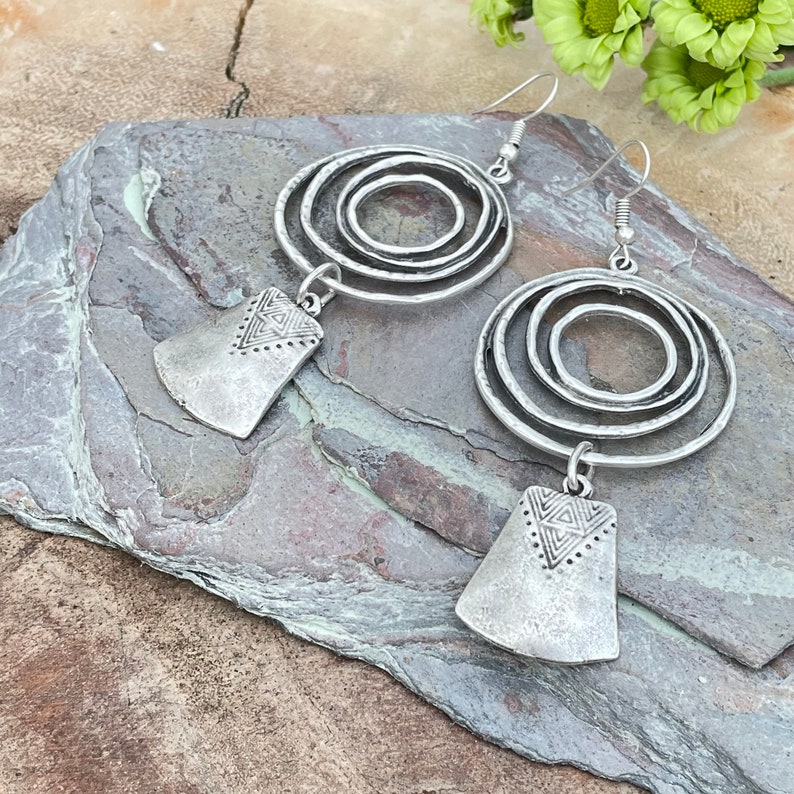 May include: A pair of silver-toned earrings. Each earring features three concentric circles and a rectangular pendant with a geometric design. The earrings have a rustic, hammered metal appearance and dangle from a simple hook.