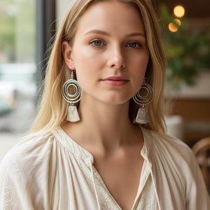 May include: Silver-toned earrings with a circular design and a rectangular pendant. The earrings are worn by a person wearing a cream-colored blouse with a V-neck and embroidered details. The background is blurred, with a window and soft lighting.