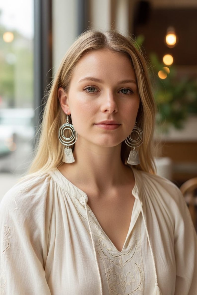 May include: Silver-toned earrings with a circular design and a rectangular pendant. The earrings are worn by a person wearing a cream-colored blouse with a V-neck and embroidered details. The background is blurred, with a window and soft lighting.