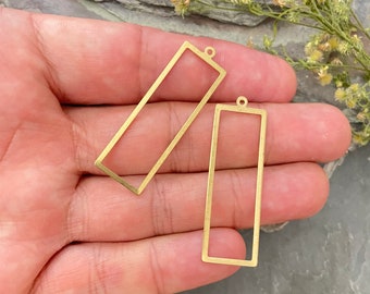 May include: Two rectangular brass jewelry charms with an open frame design. Each piece features a thin metal border and a small circular loop at the top for attachment. The finish is a matte gold tone with a smooth, flat surface texture.