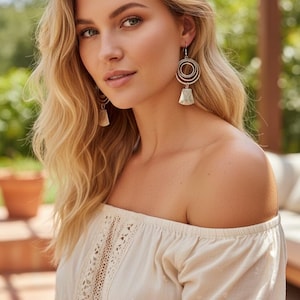 May include: A woman wearing silver earrings with a circular design and a bell-shaped charm. She is wearing an off-the-shoulder cream-colored top with lace detailing. The background is blurred, with greenery and a patio visible.