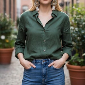 May include: A woman wearing a dark green button-down shirt and blue jeans. The shirt has long sleeves and is buttoned up. The jeans are a medium wash denim. She is wearing large hoop earrings.