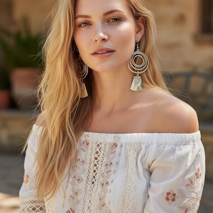 May include: A woman with long blonde hair wearing a white off-the-shoulder top with floral embroidery and lace detailing. She is wearing silver earrings with a circular design. The background is blurred, with greenery and a building visible.