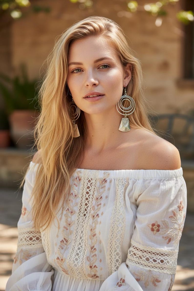 May include: A woman with long blonde hair wearing a white off-the-shoulder top with floral embroidery and lace detailing. She is wearing silver earrings with a circular design. The background is blurred, with greenery and a building visible.