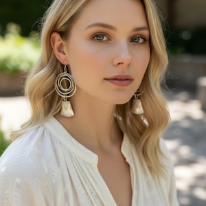 May include: Silver-toned earrings with a spiral design and a bell-shaped bottom. The earrings are worn by a person with blonde hair and a white blouse. The background is blurred with green foliage.