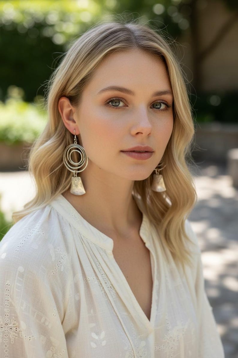 May include: Silver-toned earrings with a spiral design and a bell-shaped bottom. The earrings are worn by a person with blonde hair and a white blouse. The background is blurred with green foliage.