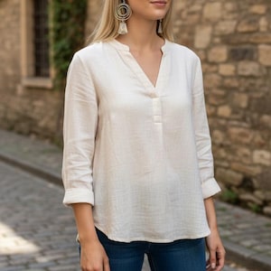May include: A woman wearing a cream-colored, long-sleeved blouse with a V-neck and rolled-up sleeves, paired with blue jeans. She is also wearing large, circular earrings. The background features a stone wall and a brick-paved street.