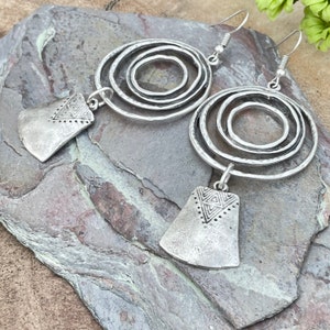 May include: Silver-toned earrings with a layered circular design and a geometric pendant. The earrings have a hammered texture and a hook closure. The earrings are displayed on a gray stone surface.