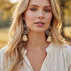 May include: A woman with long blonde hair wearing large, gold-toned earrings with concentric circles and a textured, bell-shaped bottom. She is wearing a white blouse with a delicate floral pattern.