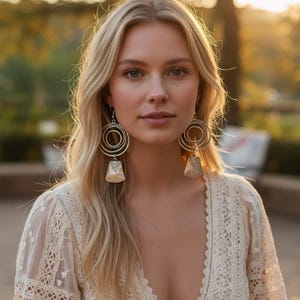 May include: A woman wearing large, silver-toned earrings with concentric circles and a bell-shaped charm. She is wearing a cream-colored lace dress with a deep V-neck. The background is blurred with warm, golden light.