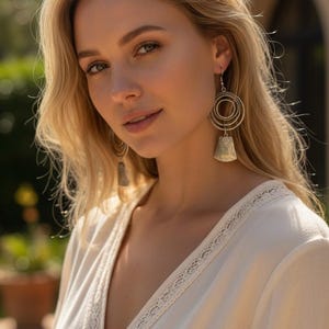 May include: A woman with blonde hair wearing silver earrings and a cream-colored top with lace trim. The earrings have a circular design with a bell-shaped pendant. The background is blurred with greenery and a building.