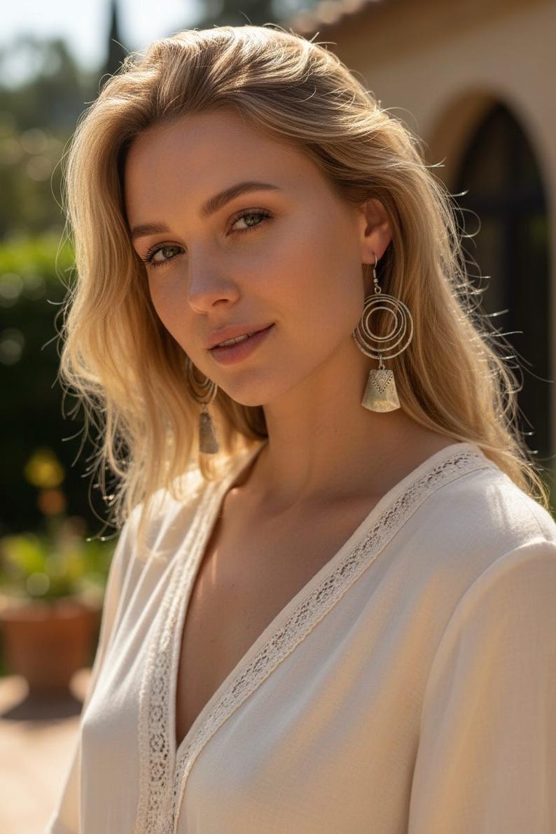 May include: A woman with blonde hair wearing silver earrings and a cream-colored top with lace trim. The earrings have a circular design with a bell-shaped pendant. The background is blurred with greenery and a building.