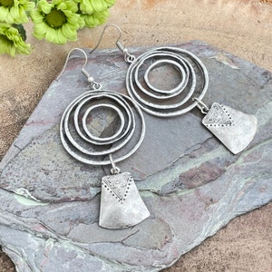 May include: Silver-toned earrings featuring concentric circles and geometric pendant drops. The earrings have a textured, antique finish and are designed with a hook closure. The earrings are displayed on a gray stone surface.