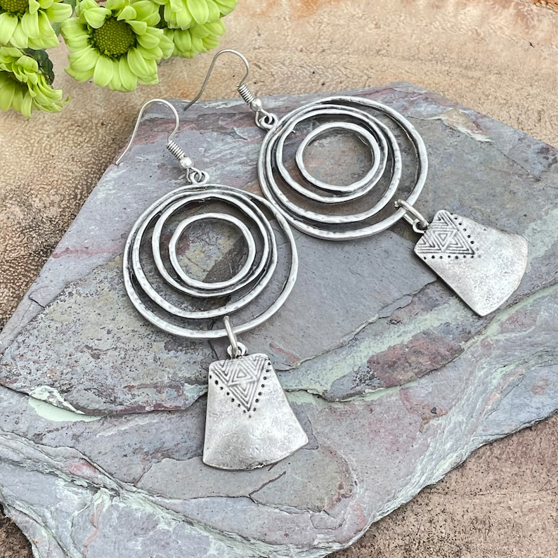 May include: Silver-toned earrings featuring concentric circles and geometric pendant drops. The earrings have a textured, antique finish and are designed with a hook closure. The earrings are displayed on a gray stone surface.