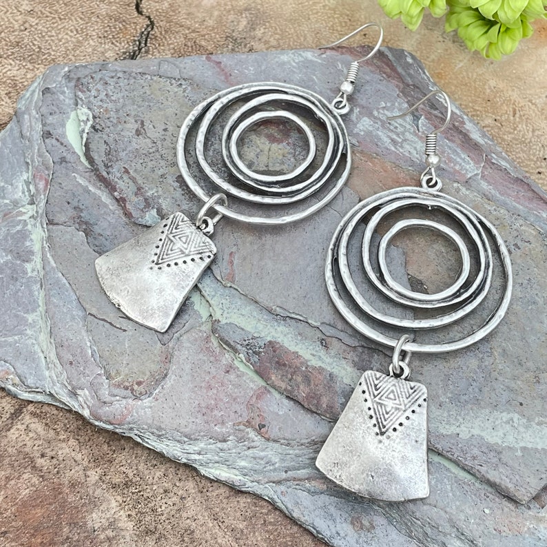May include: A pair of silver-toned earrings. Each earring features three concentric circles and a rectangular pendant with a geometric design. The earrings have a rustic, antique appearance and are displayed on a gray stone surface.