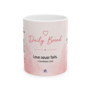 Puede incluir: Una taza de cerámica blanca con un diseño de acuarela rosa. La taza presenta las palabras "Daily Bread" en una fuente de escritura, un símbolo de corazón y la frase "Love never fails. 1 Corinthians 13:8."