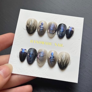 Peut inclure: Un ensemble de faux ongles ovales faits à la main. Les ongles présentent divers motifs, notamment des dégradés fumés, des illustrations de serpents et des motifs célestes dans des tons de noir, de gris et de bleu. La carte indique "HANDMADE NAIL".