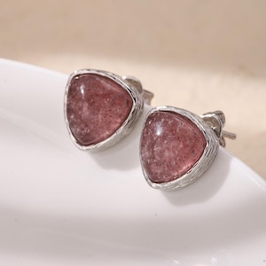 May include: A pair of stud earrings featuring triangular, pinkish-red gemstones set in silver-colored metal. The earrings are displayed on a white, curved surface, with soft lighting creating subtle shadows. The gemstones have a speckled appearance.