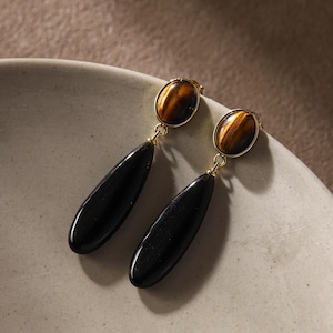 May include: A pair of elegant drop earrings. Each earring features a gold-colored top with an oval tiger's eye stone, connected to a long, teardrop-shaped black stone. The earrings are displayed on a white ceramic plate.