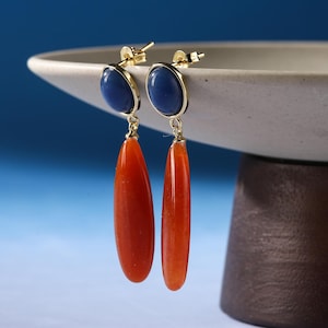 Peut inclure: Paire de boucles d'oreilles dorées avec fermoir à tige. Chaque boucle d'oreille présente une pierre ovale bleue en haut et une longue pierre orange en forme de goutte suspendue en dessous. Les boucles d'oreilles sont présentées sur fond bleu.