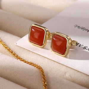Könnte beinhalten: Ein Paar quadratischer Ohrstecker mit roten Edelsteinen und goldfarbenen Metalleinfassungen. Die Ohrringe werden auf einer weißen Oberfläche präsentiert, mit einer goldenen Kette im Vordergrund. Der Text "ALWAYS AB..." ist teilweise sichtbar.