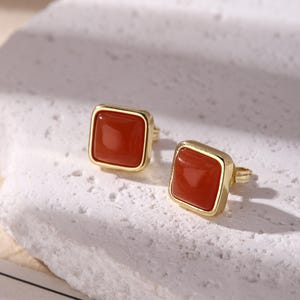 May include: A pair of square stud earrings with a gold-colored frame and a reddish-brown center. The earrings are displayed on a textured, off-white surface. The earrings are a simple, elegant design.