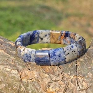 May include: A blue and white sodalite bracelet with a square bead design. The bracelet is made of natural stone and has a mix of blue, white, and brown tones. The bracelet is displayed on a tree branch.