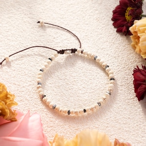 May include: A beaded bracelet featuring white and dark gray beads strung on a brown cord. The bracelet is adjustable and rests on a textured white surface, surrounded by flowers in shades of pink, yellow, and burgundy.
