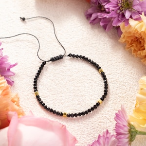 May include: A black beaded bracelet with gold-colored accents. The bracelet is adjustable with a black cord and is surrounded by colorful flowers, including pink, purple, and yellow blooms. The beads appear to be small and faceted.