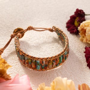 May include: A wrap bracelet featuring colorful, cylindrical beads in shades of blue, green, and orange. The beads are strung together with a gold chain and a tan leather cord. The bracelet is secured with a gold button clasp.