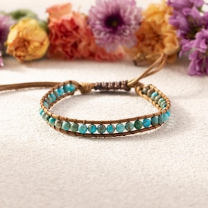 May include: A wrap bracelet with turquoise and green beads on brown leather. The bracelet is adjustable and has a braided closure. The background features colourful flowers.