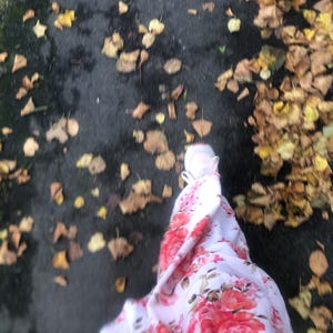 Puede incluir: Una persona con un vestido blanco con estampado floral rojo camina por un sendero cubierto de hojas caídas.