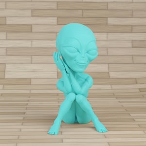 Puede incluir: Una figura de alienígena turquesa está sentada con las manos tocando suavemente su rostro. El alienígena tiene ojos grandes en forma de almendra y una ligera sonrisa. La figura está sobre una superficie de madera marrón claro.