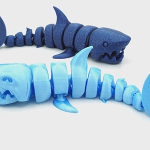 May include: Two articulated shark toys in the image. One is a dark blue color, and the other is light blue. The sharks are segmented, allowing for flexible movement. The toys have detailed features, including fins and open mouths.