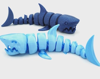 May include: Two articulated shark toys in the image. One is a dark blue color, and the other is light blue. The sharks are segmented, allowing for flexible movement. The toys have detailed features, including fins and open mouths.