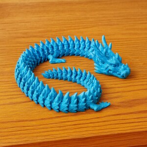 May include: A vibrant blue, articulated dragon figurine. The dragon is posed in a curved position, with a detailed head and spiky dorsal fins. The figurine is made of plastic and sits on a wooden surface.